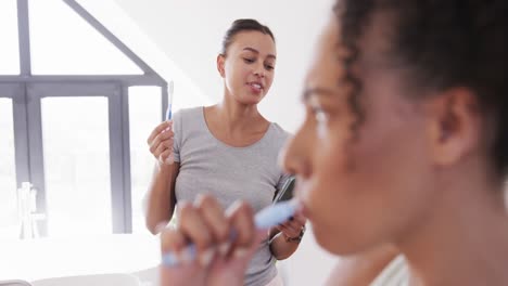 feliz pareja de lesbianas biraciales cepillándose los dientes y usando el teléfono inteligente en un baño soleado, cámara lenta