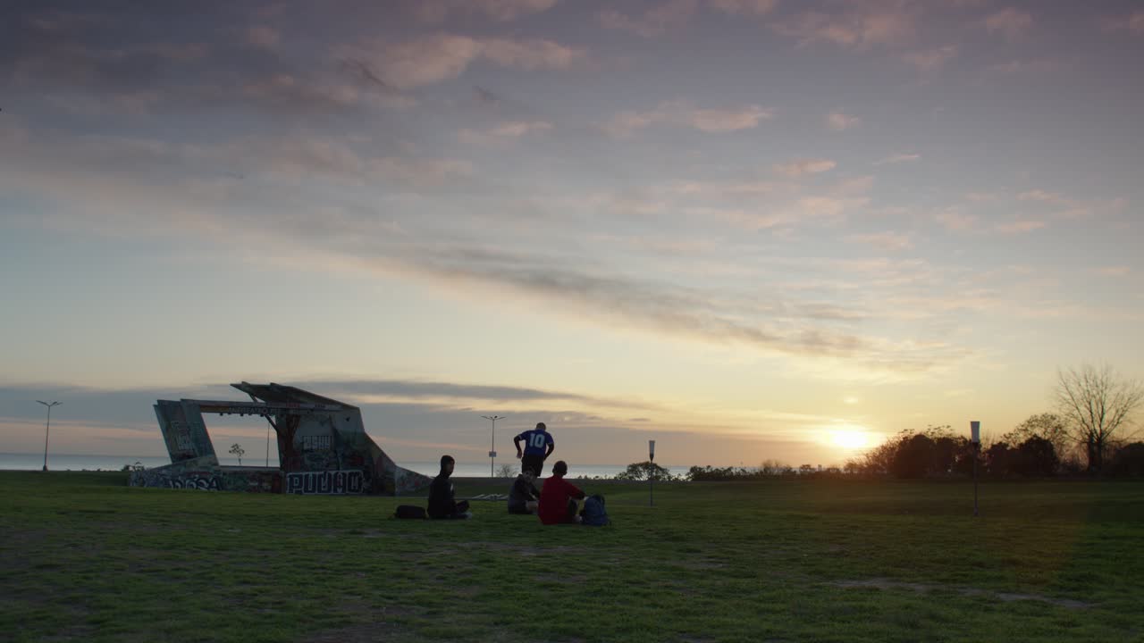 Friends play football soccer at sunset in a grassy park near graffiti-covered ruins by the sea