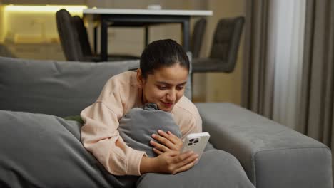 Happy-brunette-Indian-girl-in-a-beige-sweater-lies-on-a-gray-sofa-and-hugs-a-pillow-while-typing-on-a-white-phone-at-home-on-social-networks