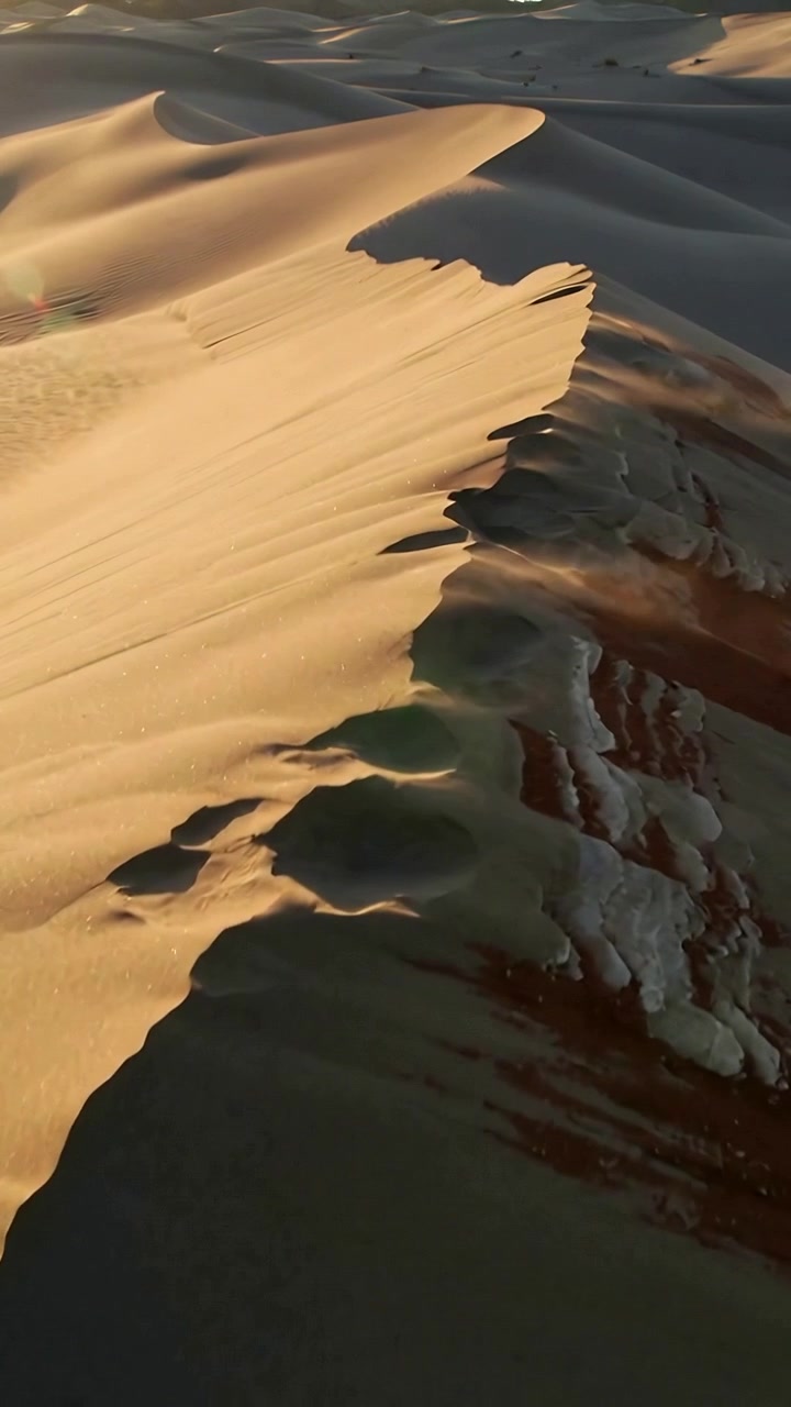 Video verticale: Drone che sorvola e cattura la cresta di una duna di sabbia all'ora d'oro, con il vento che solleva spruzzi di sabbia.