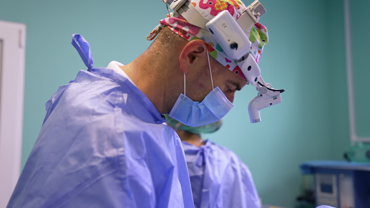 Confident male surgeon in colorful cap, mask and headlight talks to his colleague. Doctor applies tools to perform eyelid surgery. Side view.