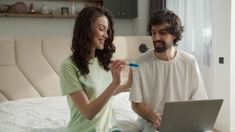 una mujer muestra una prueba de embarazo positiva a un marido sentado en la cama con una computadora portátil