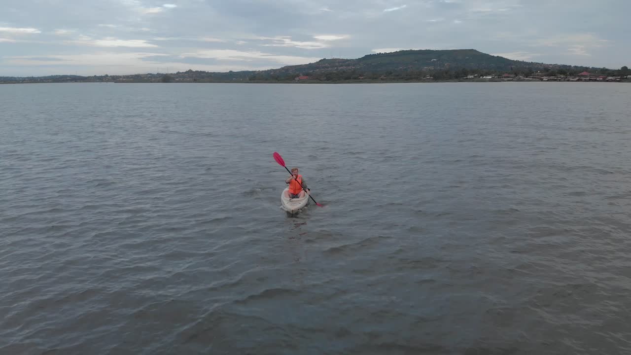 toma aérea rastreando a un hombre con un chaleco salvavidas naranja remando en el lago victoria en un kayak temprano en la mañana