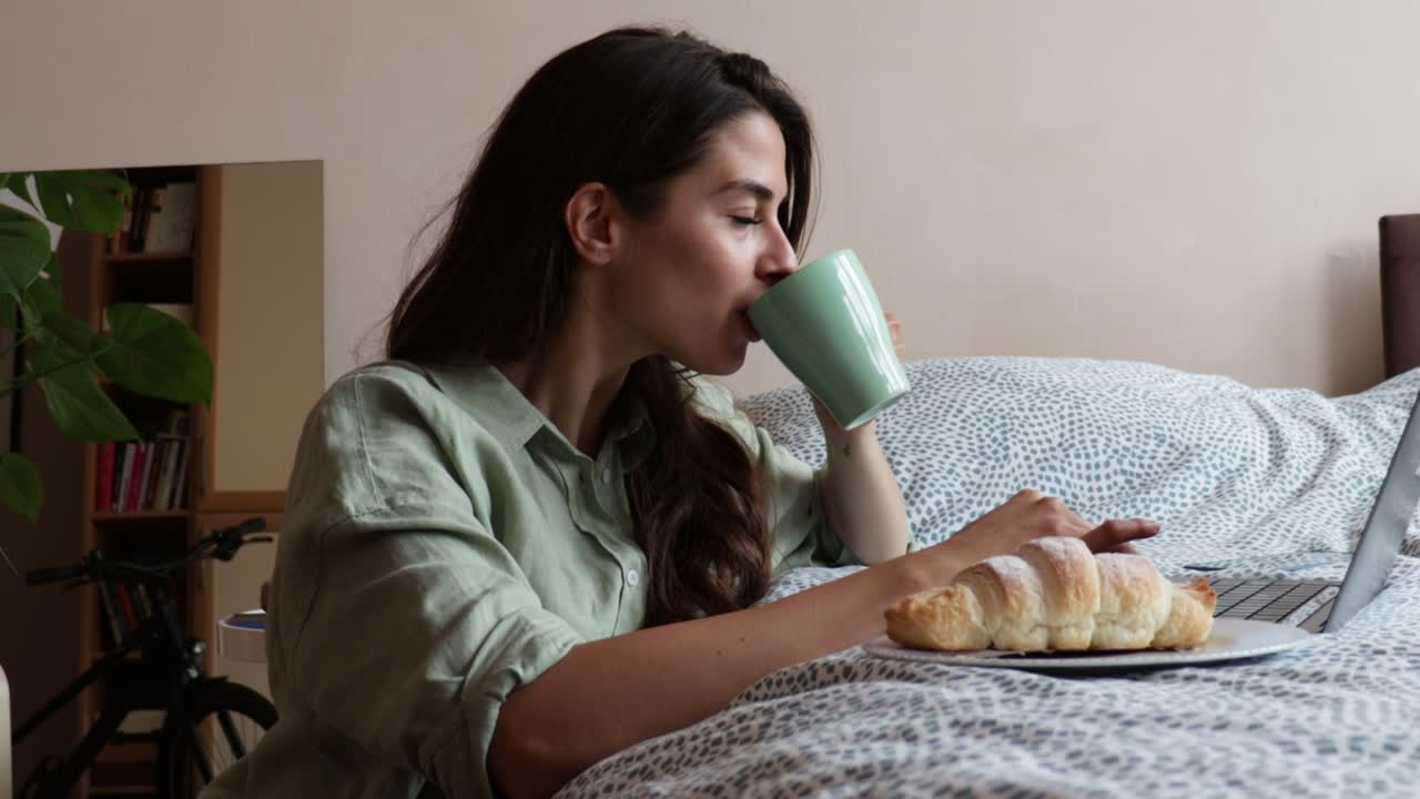 Young freelancer business woman drinking first coffee have croissant for breakfast and working on her laptop. Expatriate female using her computer to check the news in the morning at her home.