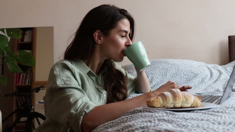 young freelancer business woman drinking first coffee have croissant for breakfast and working on her laptop. expatriate female using her computer to check the news in the morning at her home.