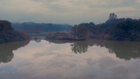 backwards drone shot of a clear reflective lake in a forest on a beautiful autumn morning in the slovenian countryside