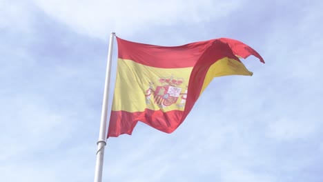 Toma-Detallada-De-La-Bandera-Española-Ondeando-En-El-Viento-En-Cámara-Lenta-Con-Cielo-Azul-Y-Nubes-En-El-Fondo