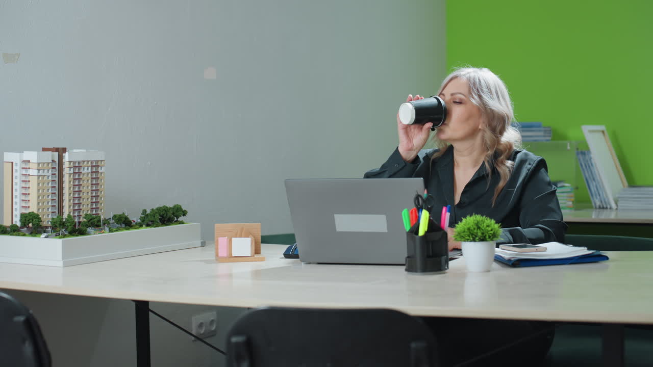 Professional saleswoman pauses work at office desk to sip coffee before resuming typing on laptop, surrounded by building model, folders, and indoor plants in modern workspace