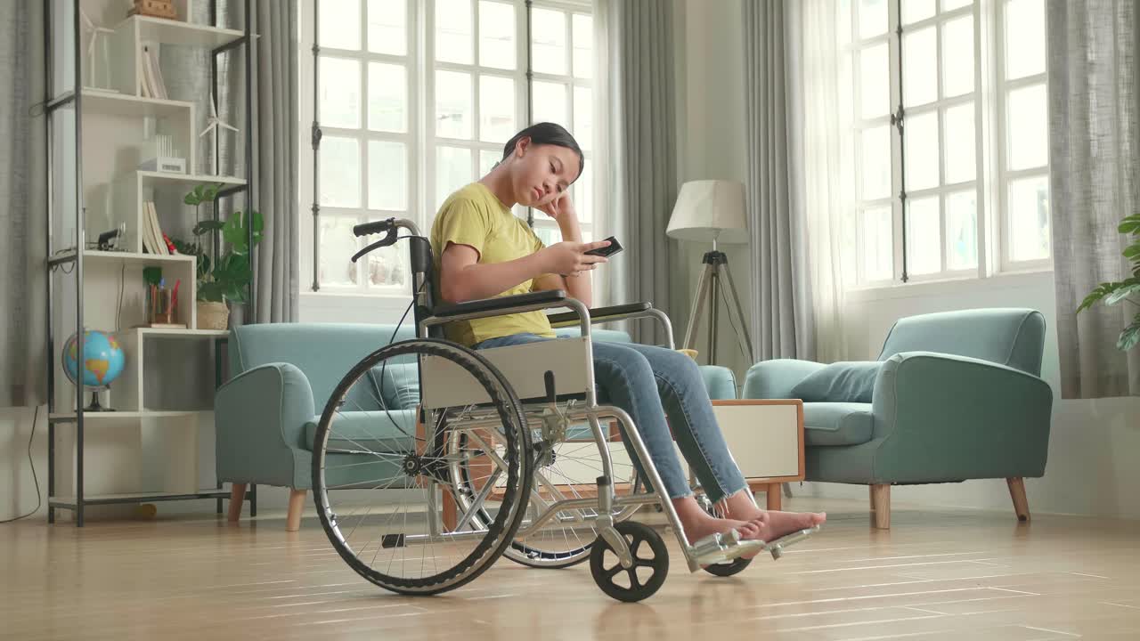Bored Asian Kid Girl Sitting In A Wheelchair While Using Smart Phone At Home