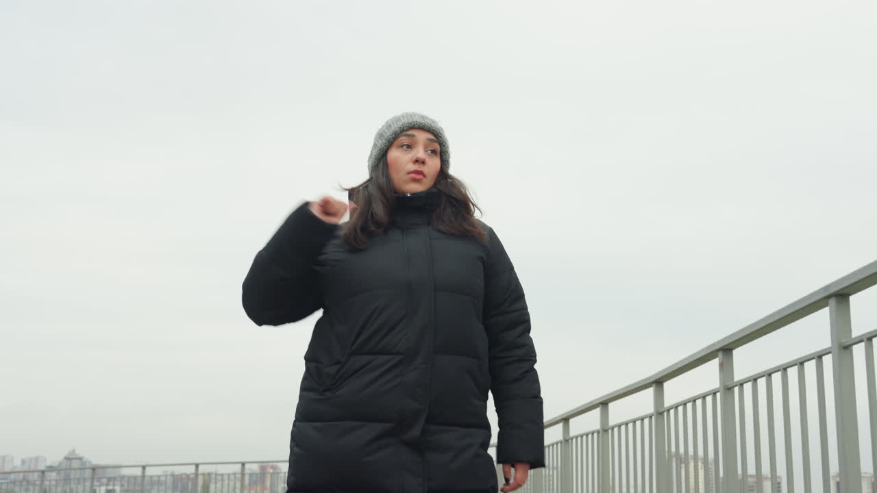 Woman dancing on city hilltop bundled in black coat and beanie, improvising expressive arm gestures along railing, alternating wide steps and close portraits, overcast skyline backdrop, cold breath