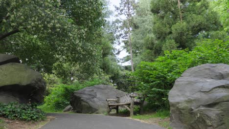 Wenn-Man-Einen-Naturpfad-Aus-Schwarzem-Asphalt-Hinaufsteigt,-Entdeckt-Man-Beim-Erreichen-Der-Hügelkuppe-Grüne-Bäume,-Büsche,-Einen-Großen-Felsblock-Und-Eine-Rustikale-Blockparkbank