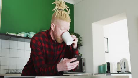 hombre albino afroamericano pensativo con rastas usando un teléfono inteligente y bebiendo café