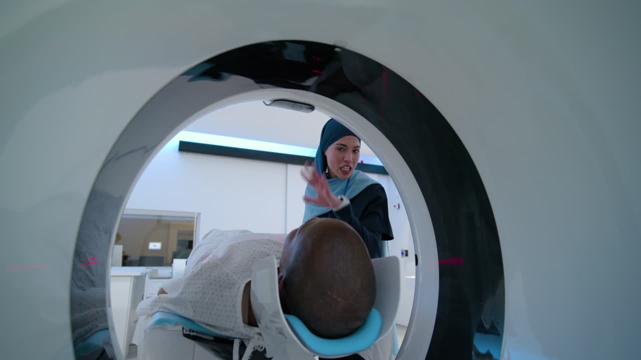 Tecnico con hijab e paziente maschio nella sala scanner, il tecnico regola il cuscino blu e conferma l'avvio della scansione