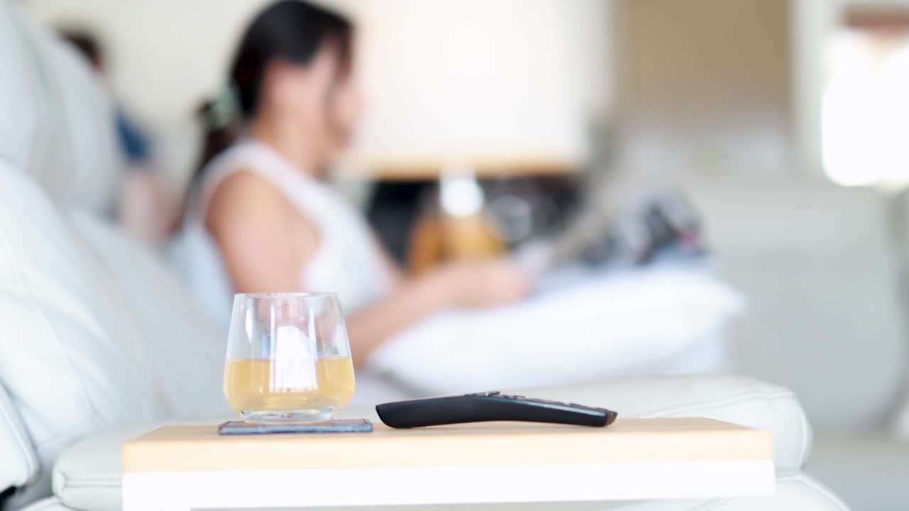 A woman sits on a modern sofa in a bright living room, reading a magazine. A remote control and drink rest on a nearby table. Shallow depth of field, static camera