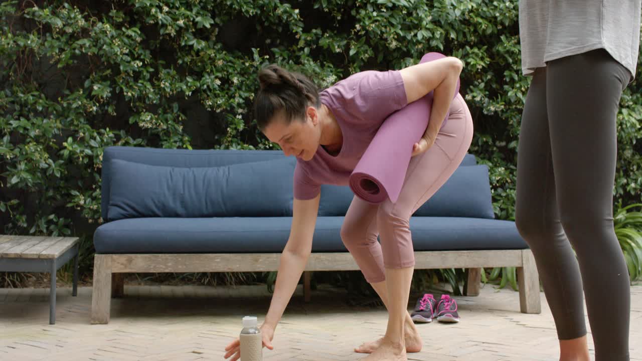 Diverse female friends arriving, preparing yoga mats in activewear on patio placing water bottles