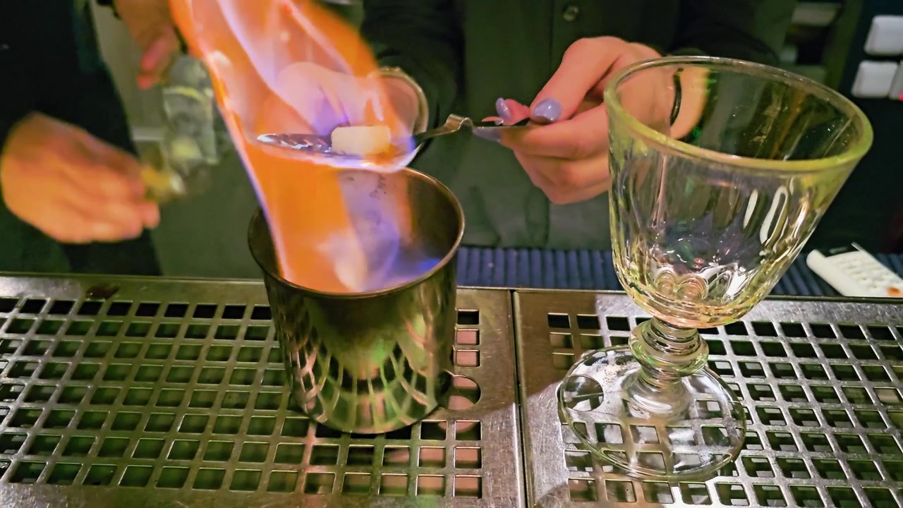 Primo piano di un cocktail scenografico in preparazione con fiamma blu in un bar, che mostra una zolletta di zucchero, un cucchiaio forato, una tazza di metallo, un calice vuoto, le mani del barista e un'atmosfera da lounge scura.