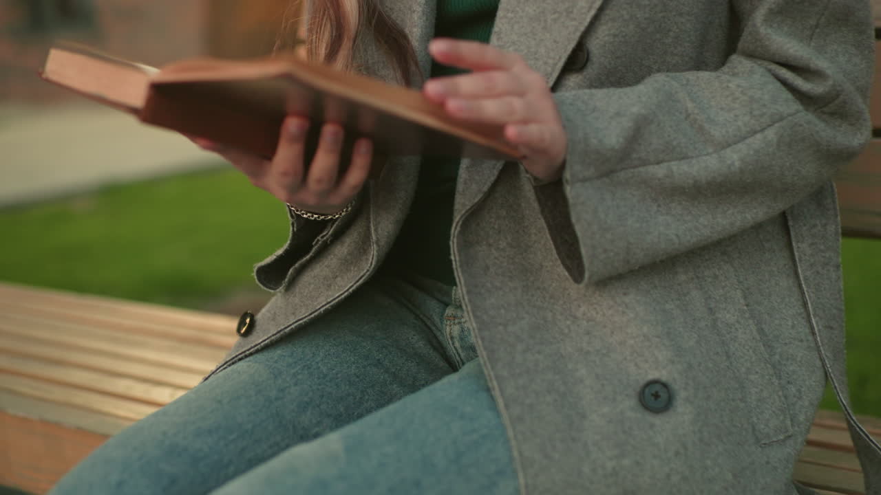 Close up of woman seated on wooden bench with open book in hand, wearing grey coat and denim jeans, casual outdoor setting with focus on relaxed posture and study moment in natural environment