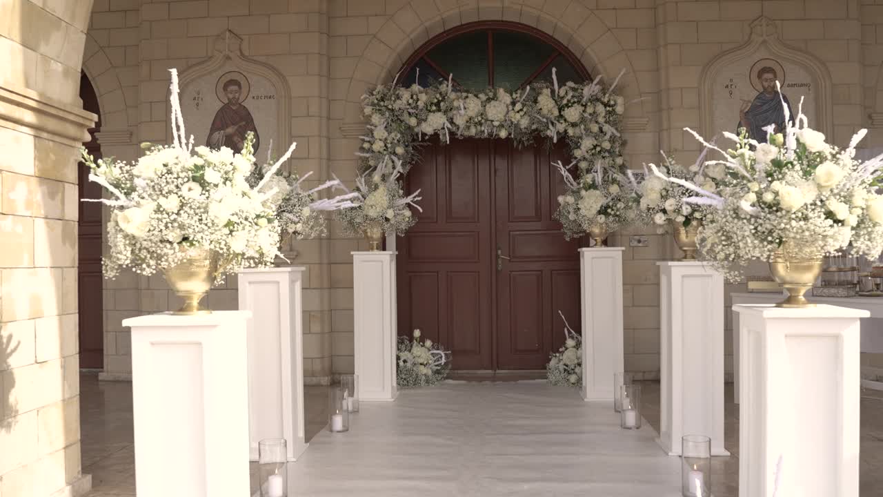 Composizioni floreali decorano l'ingresso di una chiesa in un paesino. Un tappeto bianco conduce alle porte, creando uno spazio invitante per gli ospiti. L'evento si svolge nel pomeriggio.
