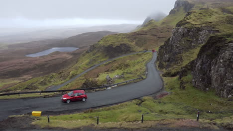 panning shot from a vista of rocky cliffs, meadows and lakes with curvy road in the foreground with passing cars on a rainy and cloudy day in quiraing, scotland, isle of skye