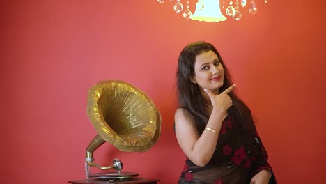 An-Indian-woman-in-black-saree-standing-next-to-gramophone-smiling-at-the-camera-in-an-isolated-red-background-with-golden-light-on-top