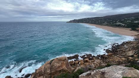 Slow-motion-of-coastal-sea-waters,-waves-are-crashing-at-shore-banks,-in-distance-visible-residential-area-with-houses