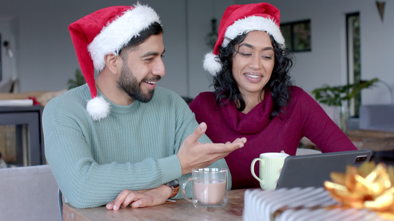 una feliz pareja biracial con sombreros de papá noel usando una tableta para una llamada de video en casa, en cámara lenta