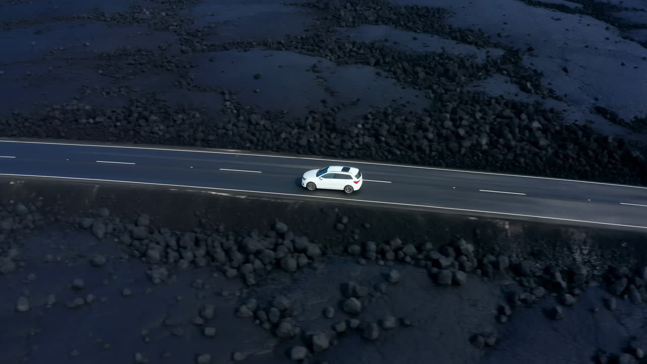 火山性の景観の中を道路を走る白い車の空撮。
