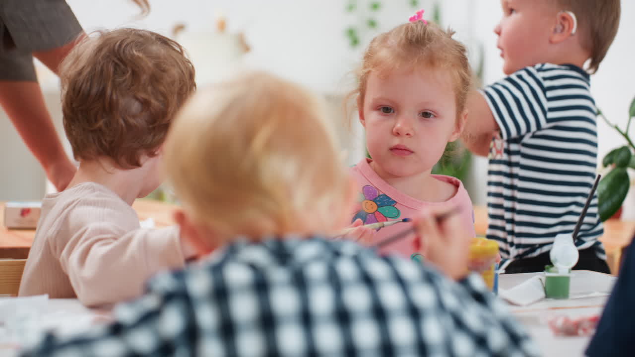Close up of cute children engaged in painting session, with special child wearing hearing aid holding brush showing his work to instructor while classmates focus on creative activity at table indoors