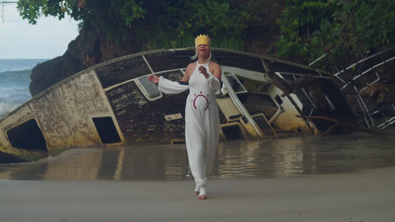 A wide shot of a girl in costume walking on the wet sand, her reflection visible in the water under a moody Caribbean sky