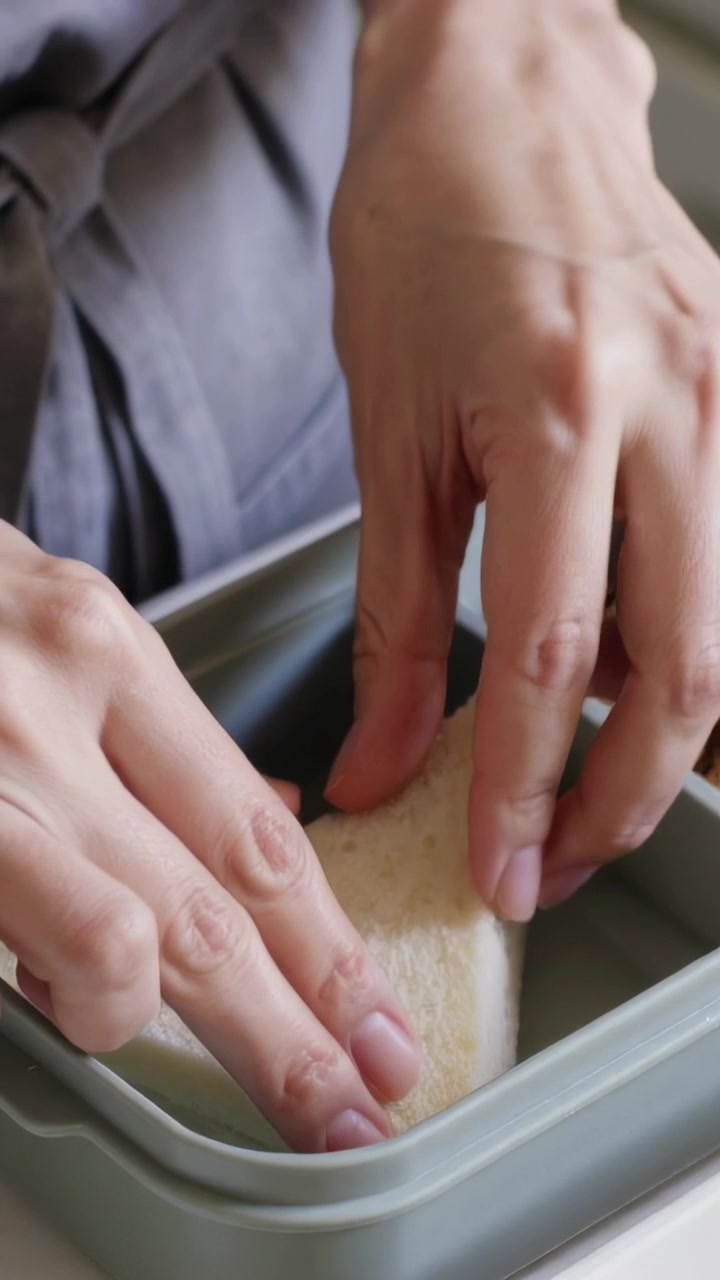 Video verticale: Mani con grembiule grigio che confezionano un sandwich bianco triangolare, inserendolo in una scatola con snack.