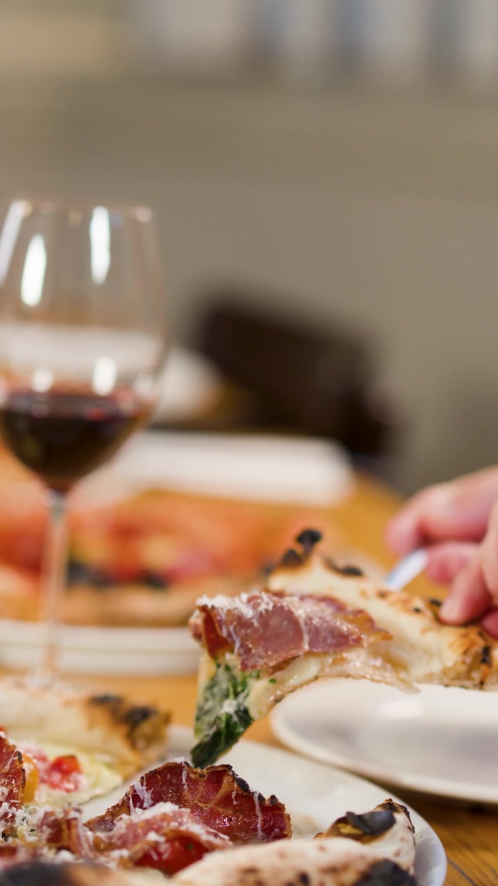 Primo piano di mani che servono una fetta di pizza artigianale su un piatto con vino rosso.
