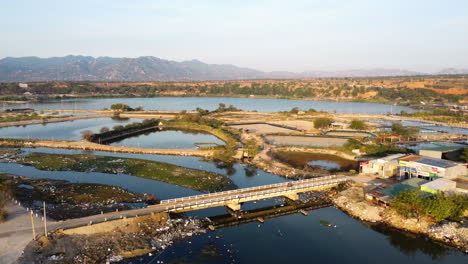 Vista-Aérea-De-La-Isla-Contaminada-Y-El-Río-De-Son-Hai,-Vietnam-Durante-El-Día-Soleado---Camiones-Cruzando-El-Puente-Y-Las-Montañas-En-El-Fondo