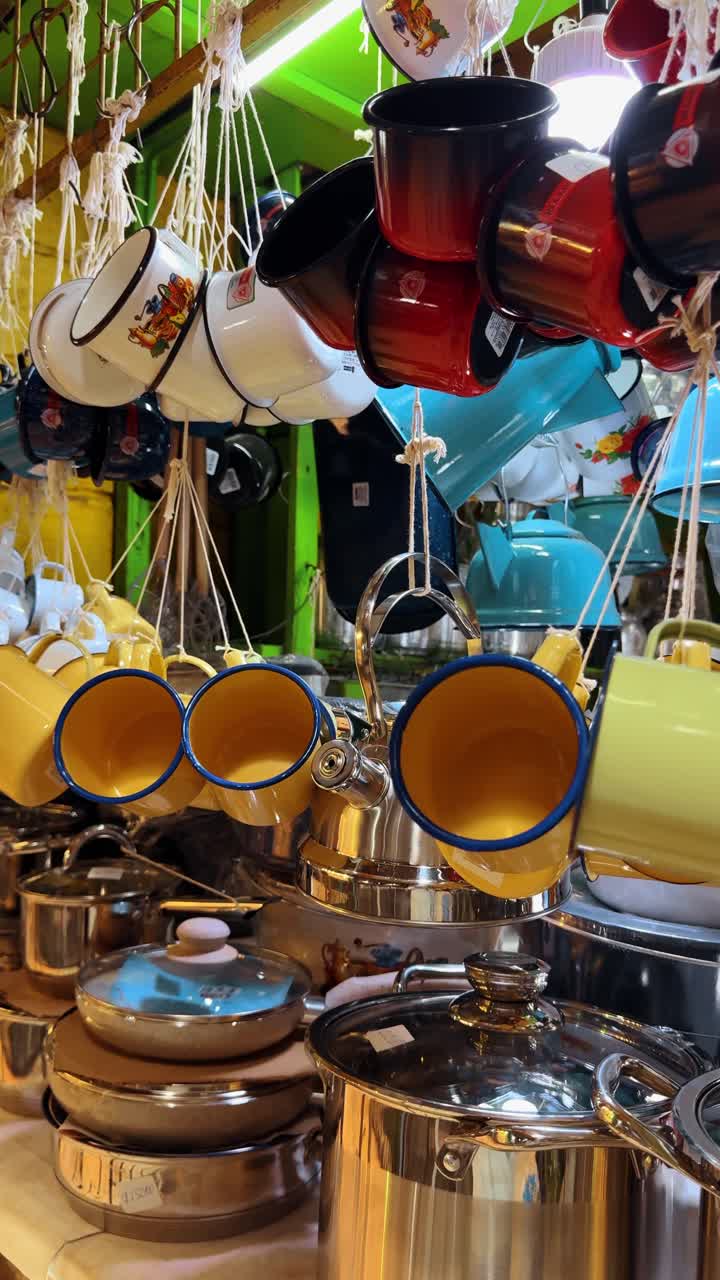 San Jose, Kosta Rika, 7 Agustus 2025: Mug enamel berwarna-warni dan peralatan masak logam tergantung di pasar lokal. Pajangan ritel mug kuning dan biru cerah dengan panci baja tahan karat di sebuah toko Kosta Rika.
