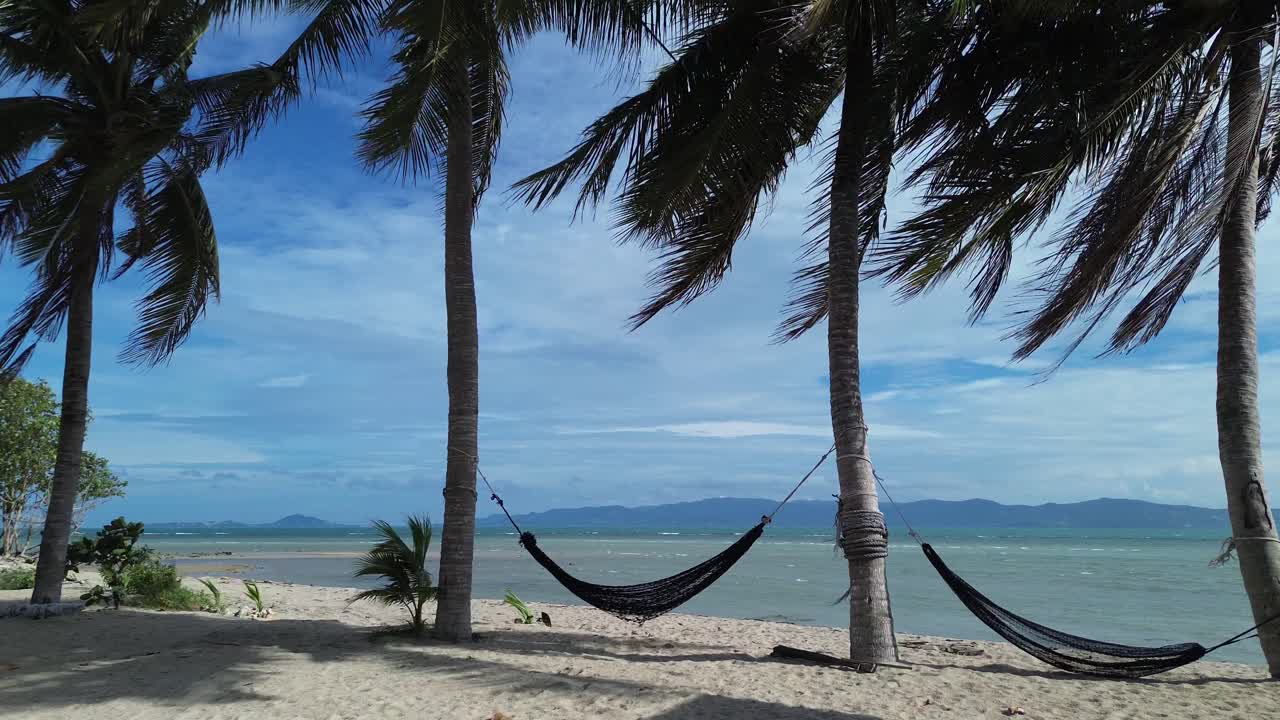 Drone footage capturing hammocks strung between palm trees on a sandy beach in Koh Phangan Thailand with turquoise sea, tropical coastline, and tranquil summer atmosphere