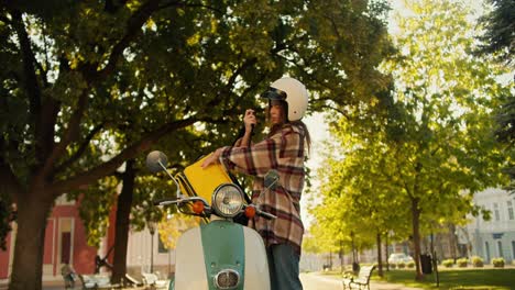 A-brunette-courier-girl-in-a-checkered-shirt-wearing-a-white-motorcycle-helmet-and-with-a-yellow-bag-comes-up-and-puts-it-on-her-moped.-A-girl-inspects-an-order-that-she-is-going-to-deliver-on-the-street-in-the-summer