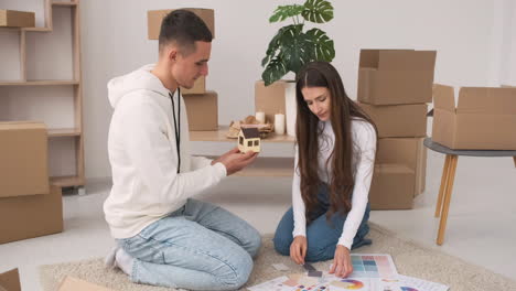 front view of a young couple in a new house sitting on the carpet and choosing colours for decoration wth a house model