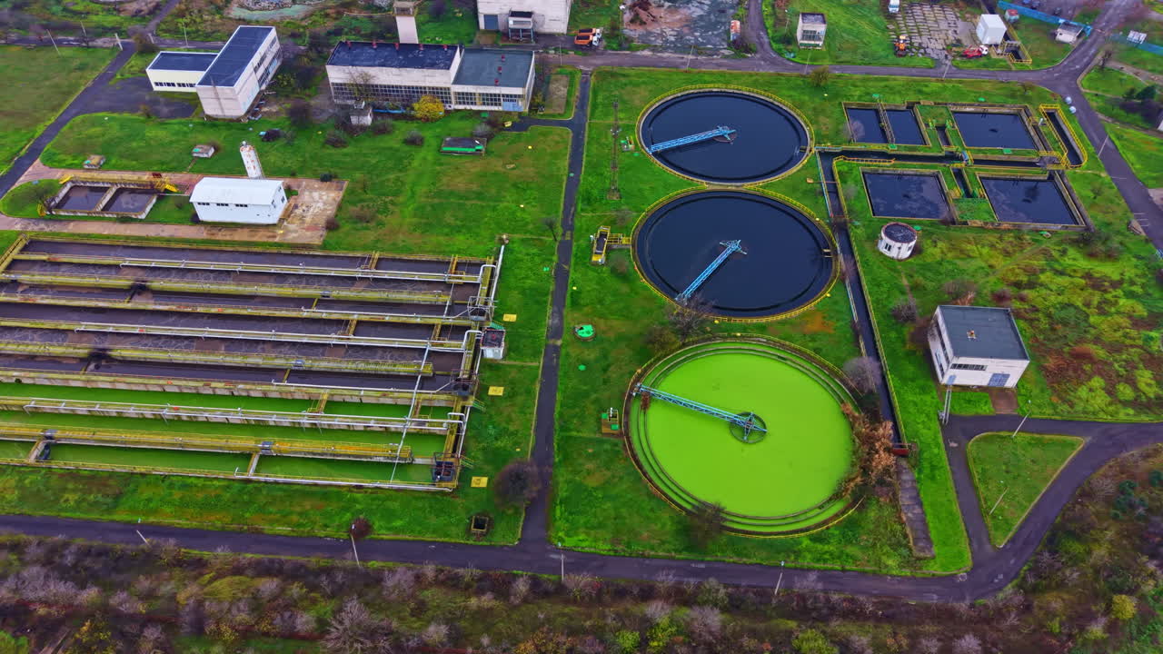 Aerial view of a wastewater treatment facility with several large tanks and various structures in a green landscape during daytime