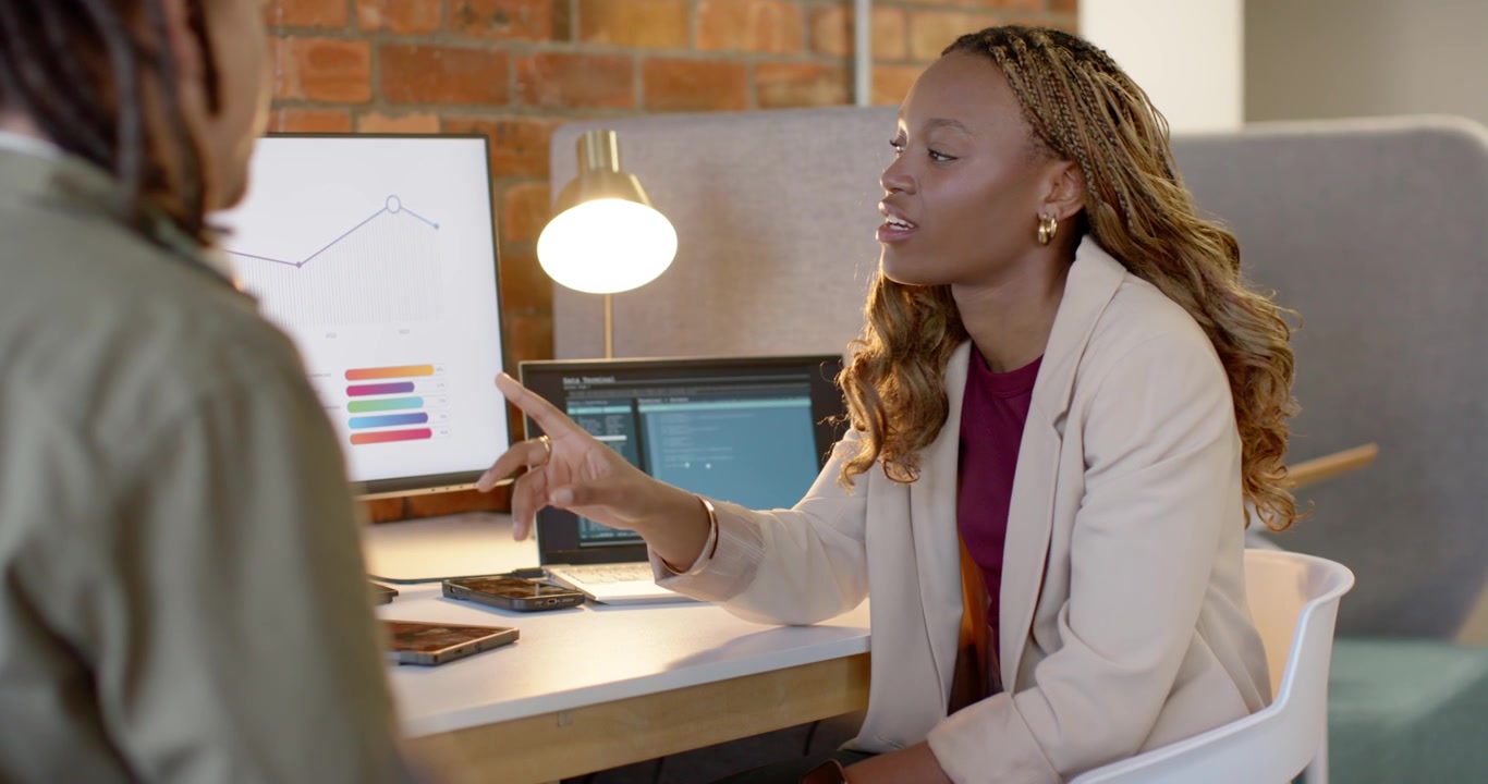 African American team colleague gesturing, seated woman at desk pointing monitor clarifying chart
