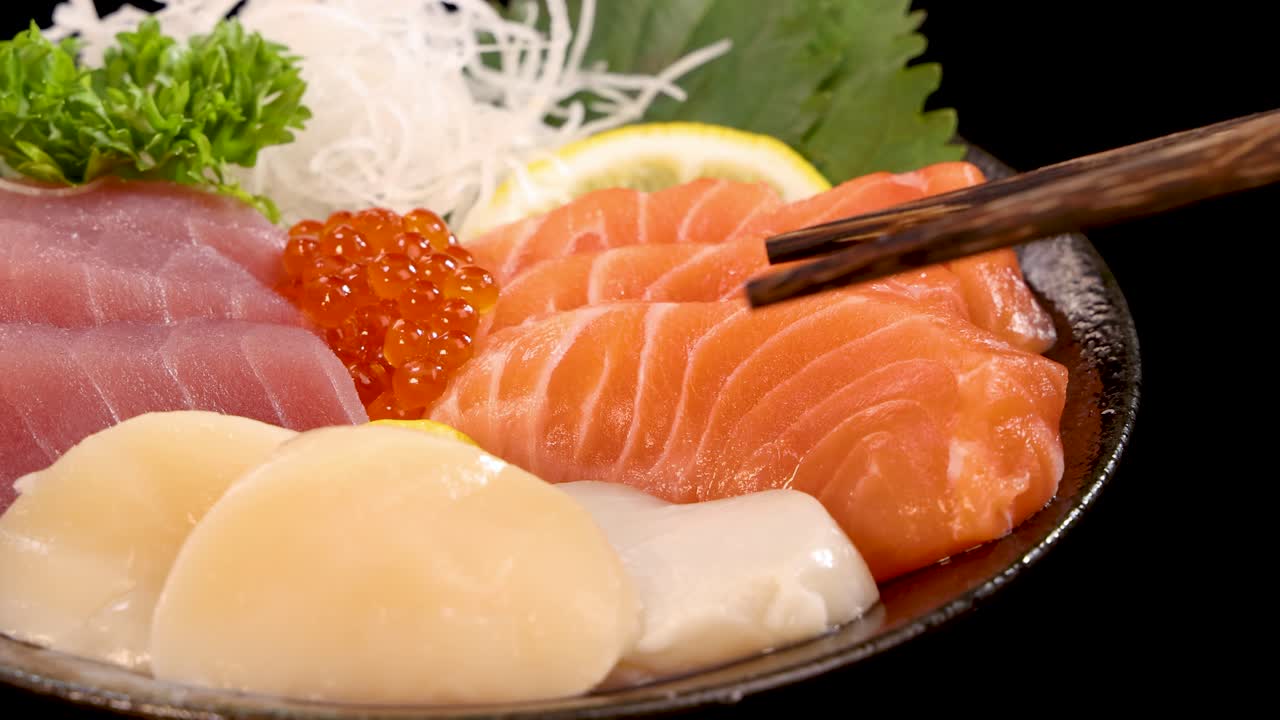 Chopsticks lift a fresh scallop sashimi from a Japanese sushi set with salmon, tuna, and roe, under bright studio lighting on a black background