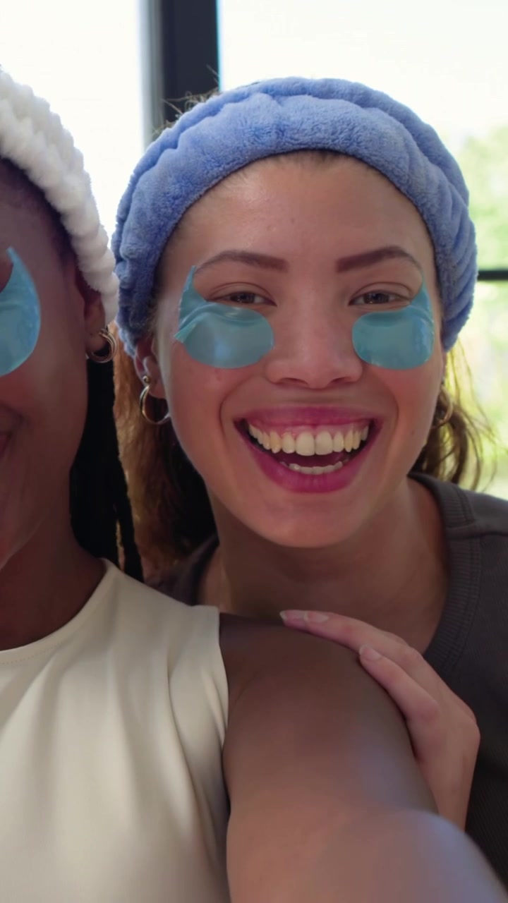 Vertical video: Smiling two women holding phone filming selfie at window showing turquoise patches