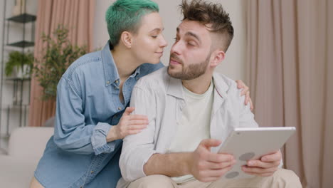 Handsome-Man-Sitting-On-Sofa-And-Holding-A-Tablet-While-His-Girlfriend-Approaching-And-Hugging-Him-From-The-Back