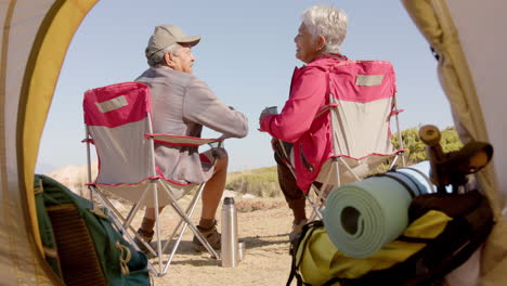 happy senior biracial couple sitting at tent in mountains and kissing, in slow motion