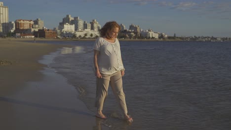 Tracking-shot-of-relaxing-pregnant-red-haired-woman-enjoying-ocean-view-and-sunlight-at-beach-during-windy-day-at-sunset