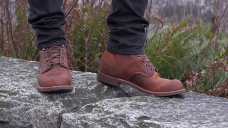 Close-up-shot-of-young-man-wearing-dark-grey-jeans-and-brown-leather-boots-standing-on-a-ledge-with-shrubs-in-the-background