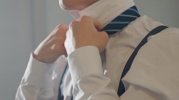 Dirigente asiatico si stringe la cravatta prima di un colloquio, camicia bianca con bretelle, mani concentrate che aggiustano il colletto, luce fredda dell'ufficio, sottile energia nervosa, ultimi momenti prima della presentazione