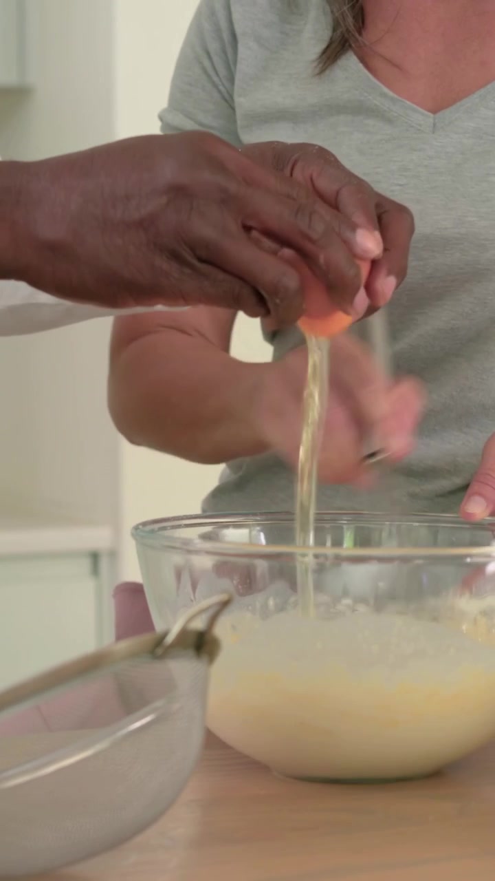 Video vertical: Mujer con camiseta gris vertiendo leche y batiendo la mezcla en la encimera de la cocina, mientras un hombre rompe huevos.