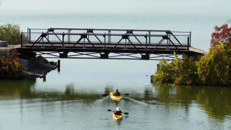 an aerial view of two people slowly kayaking away from the camera and going under a pedestrian bridge