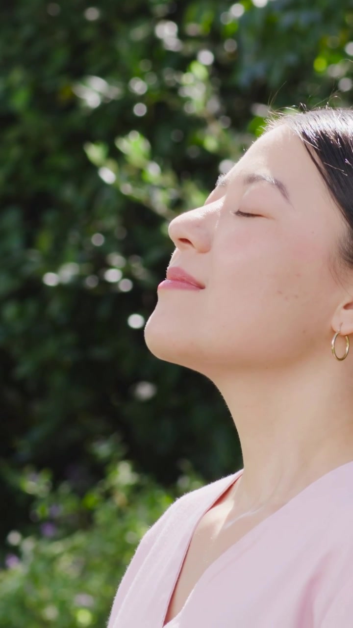 Video verticale: Luce solare che accarezza una donna con orecchini a cerchio d'oro e top rosa, che inclina la testa in giardino, con spazio per il testo.