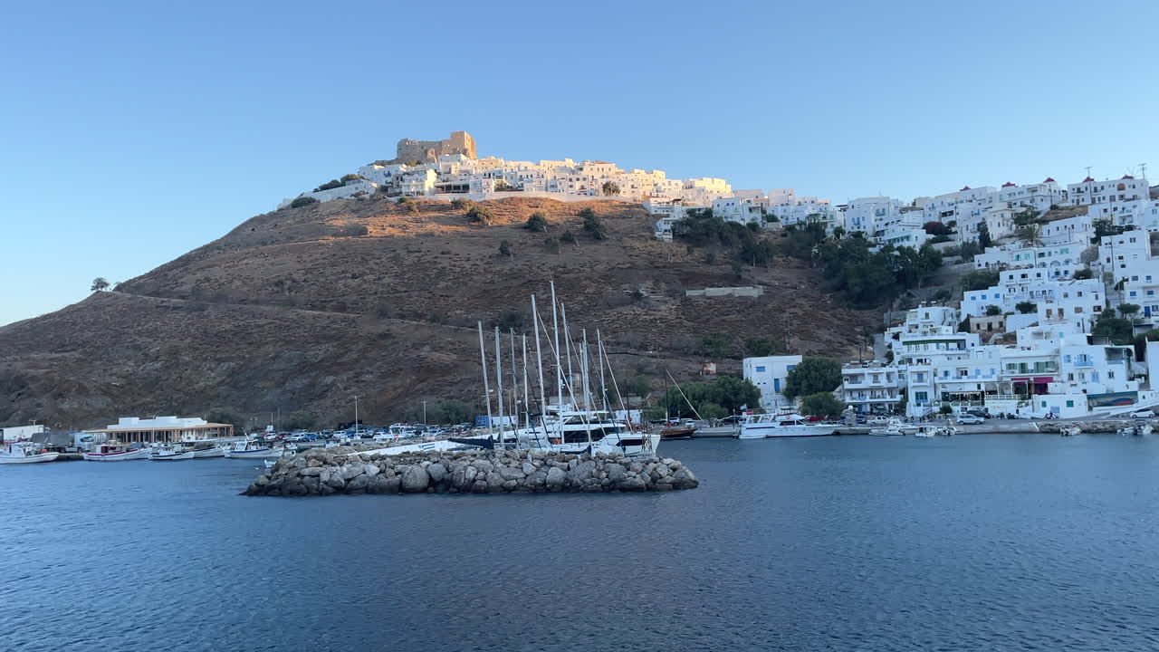Greece, Astypalea Island, footage starts from the harbor full with white beautiful sailboats and zooms towards to the castle shinning under the sun with white houses around it during sunset.