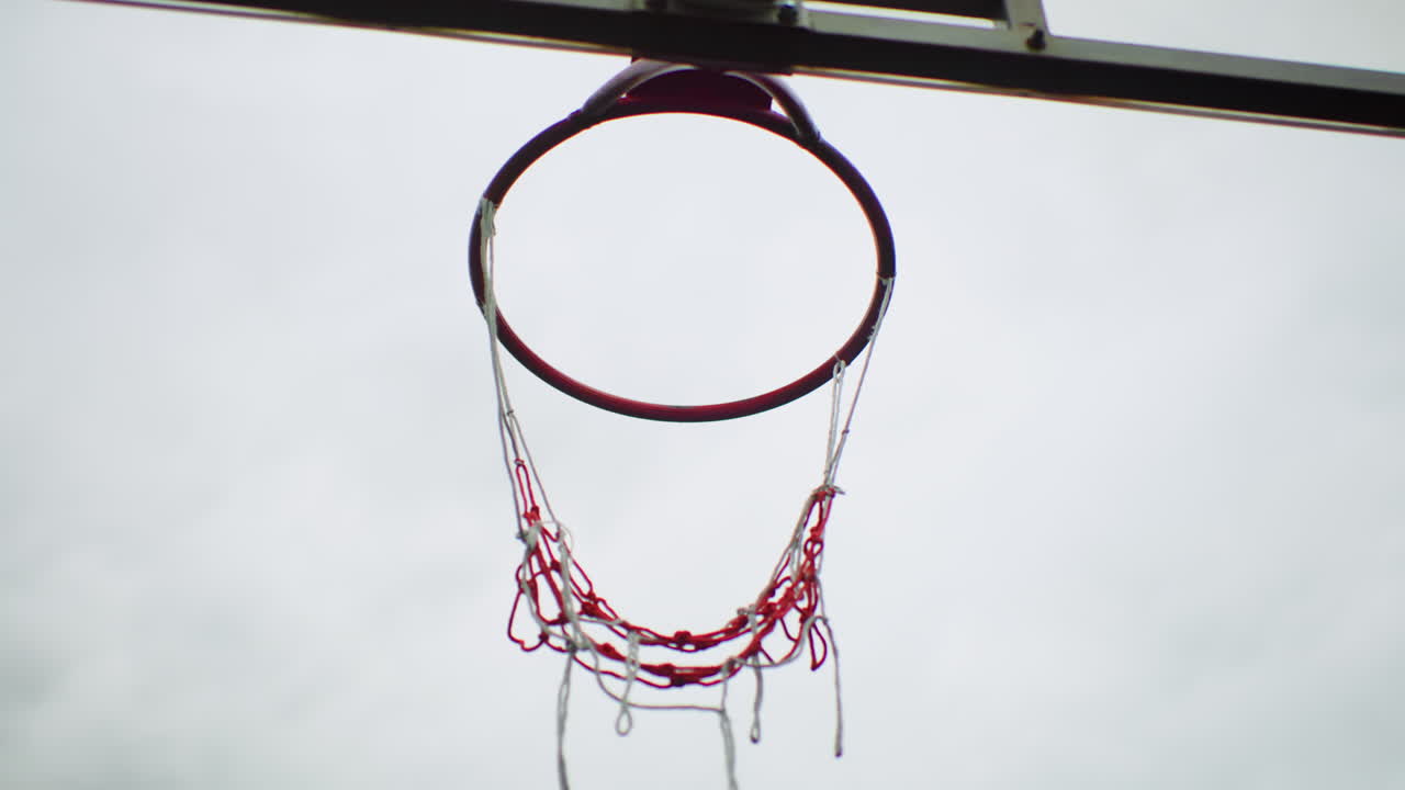 basketball hoop overcast sky swish net close underside perspective showing frayed red net swinging from metal rim, subtle motion and droplet detail, cinematic moody atmosphere of empty urban